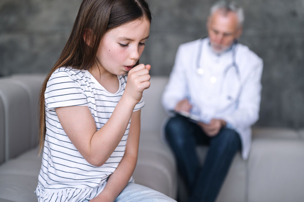 A sick child coughing while sitting on the sofa with a doctor in the background