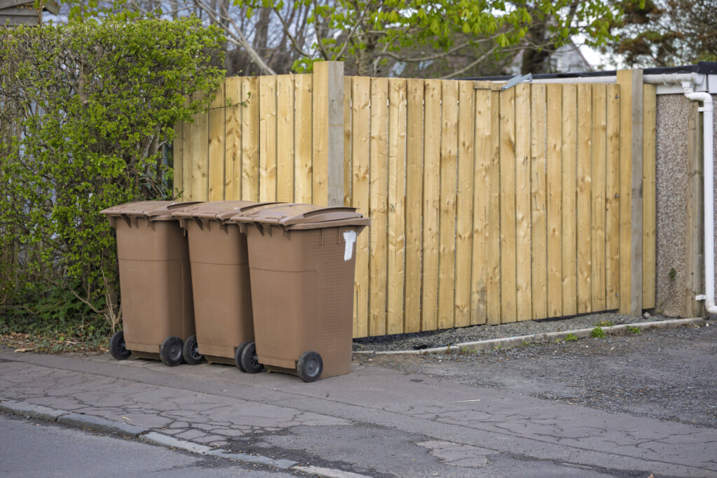 Brown wheelie bins on pavement beside wooden fence