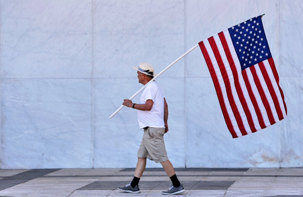 A man holds American flag as the wall of the John F. Kennedy Center for the Performing Arts is covered in tarp after President Donald Trump's name was removed, in Washington, Saturday, June 13, 2026. (AP Photo/Rahmat Gul)