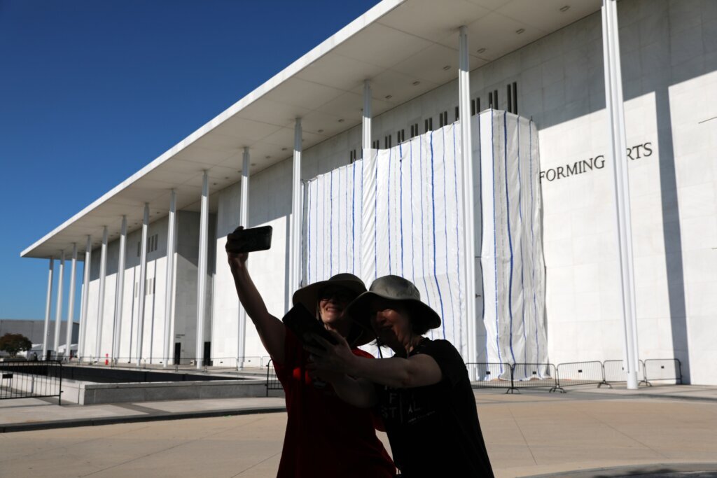 Women take a selfie as the wall of the John F. Kennedy Center for the Performing Arts is covered in tarp as work continues on the removal of President Donald Trump's name, Saturday, June 13, 2026 in Washington. (AP Photo/Rahmat Gul)