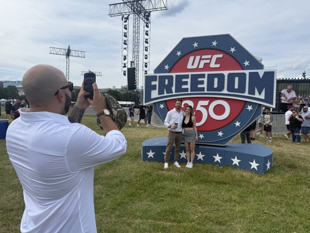 Two people taking a photo in front of the "UFC Freedom 250" sign. (WTOP/Jimmy Alexander)