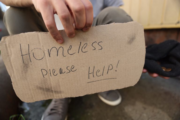 Man's hand holding cardboard sign: "Homeless please help"